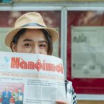 A woman wearing a fedora holding a Vietnamese newspaper outside a newsstand.